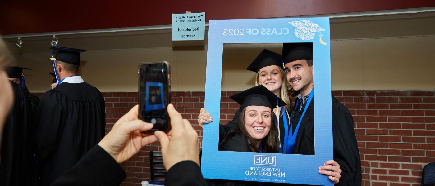 Students dressed in cap and gown pose for a photo with an oversized frame